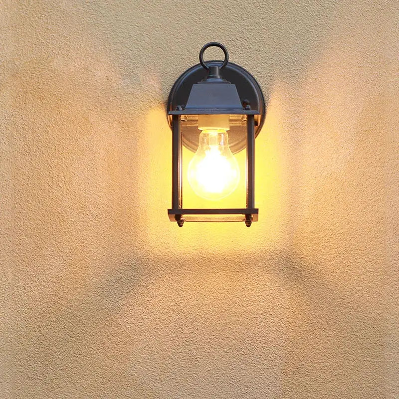 Black aluminium outdoor wall light with clear glass E27, glowing warmly on beige textured wall.