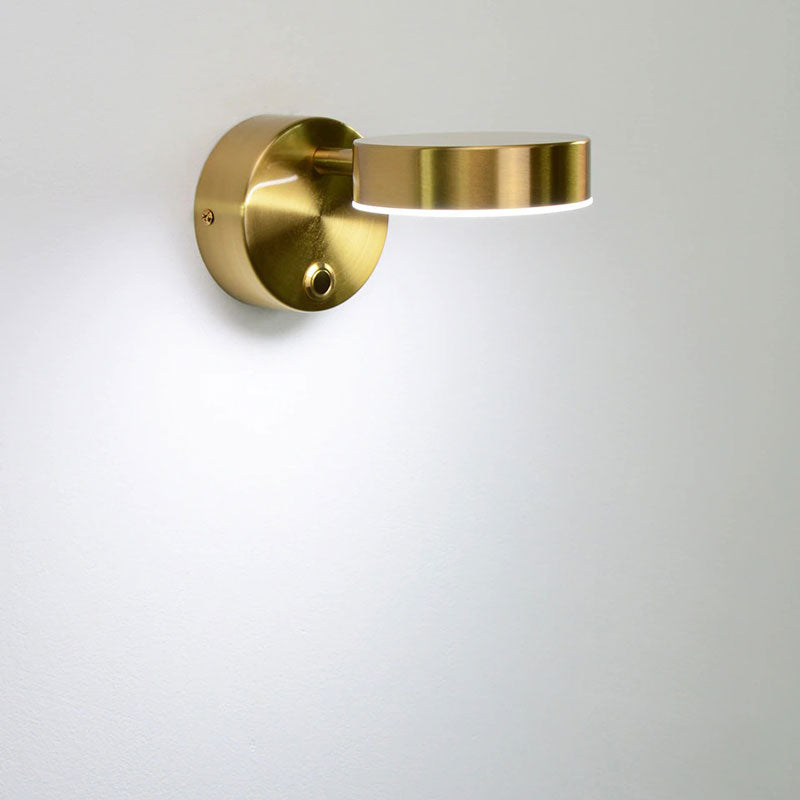 Circular brass LED wall light with flat arm and adjustable head emits soft focused glow.