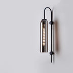Cylindrical metallic shade wall light with exposed filament bulb on minimalist black fixture.