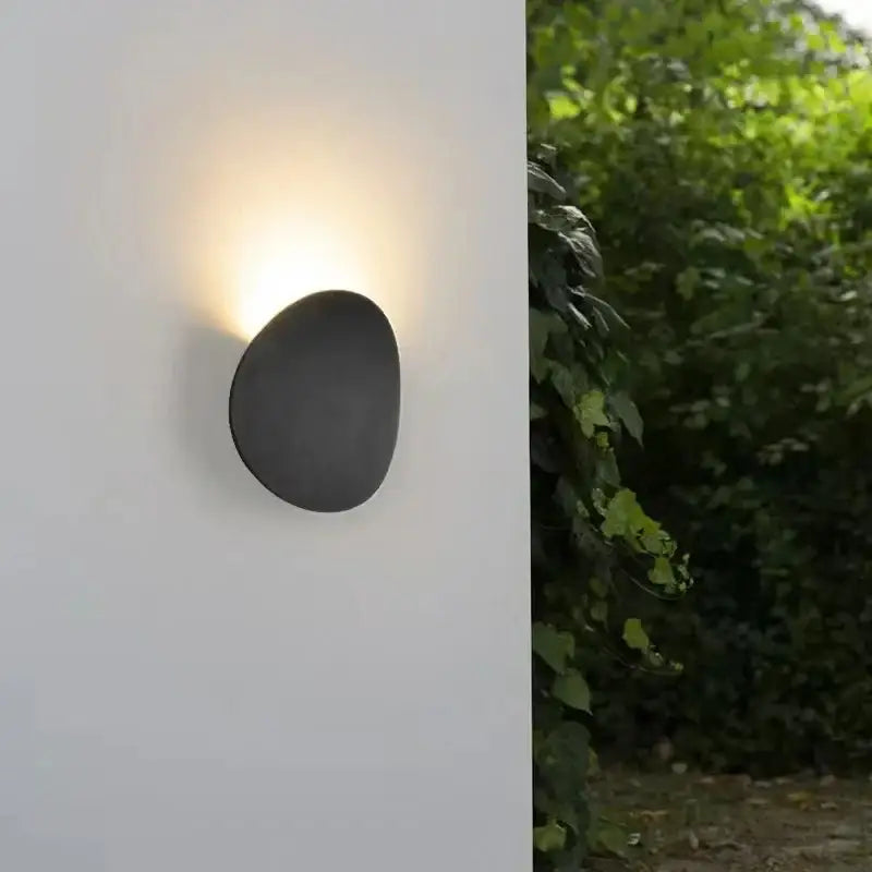 Modern black, pebble-shaped aluminum LED wall light with warm upward glow on white wall.