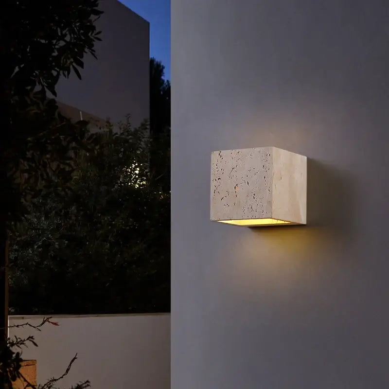 Modern cube wall sconce with a stone-textured pumice finish, glowing warmly at dusk.