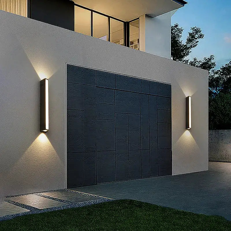 Rectangular aluminum outdoor LED wall light installed beside a garage, illuminated at dusk.
