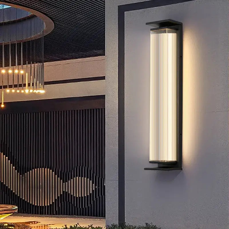 The Original Large Black Glass Outdoor Wall Light with vertical lines, softly illuminated at night.