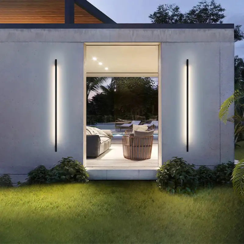 Two Slim Outdoor LED Wall Lights in Aluminum flank a wide doorway, glowing warmly at dusk.
