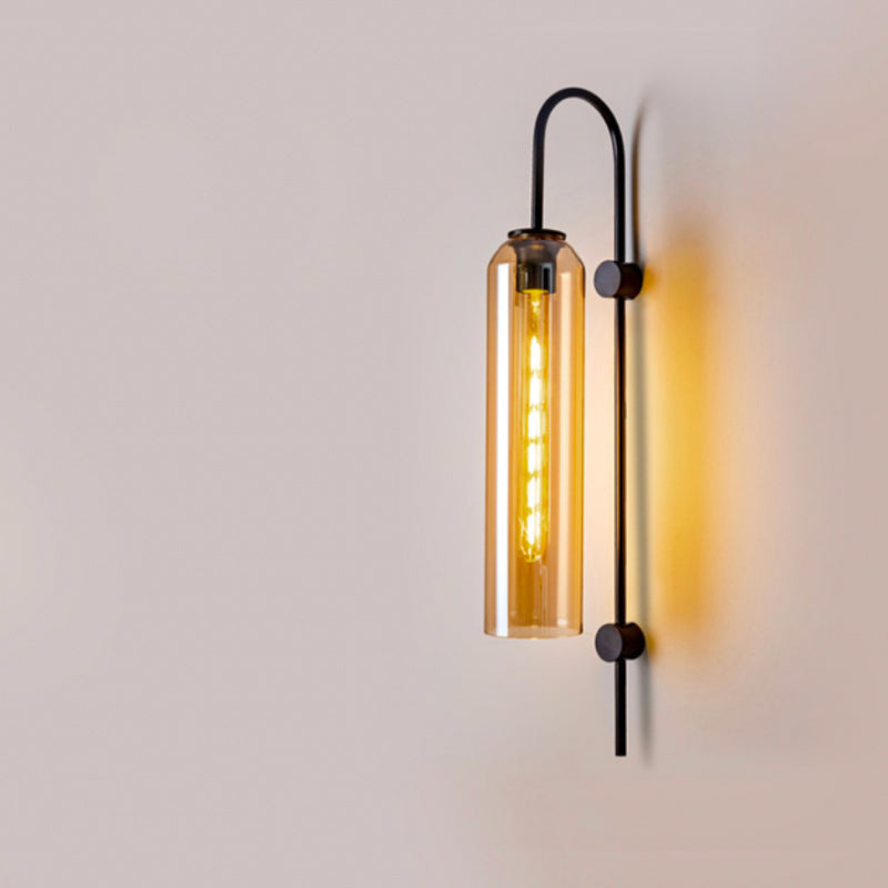 Vertical cylindrical glass wall light with exposed bulb and sleek metal bracket, glowing warmly.