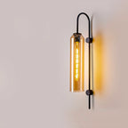 Vertical cylindrical glass wall light with exposed bulb and sleek metal bracket, glowing warmly.