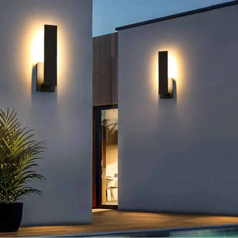 Luminaire extérieur mural moderne en aluminium noir diffusant une lumière chaude au crépuscule.