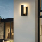 Luminaire mural en aluminium noir moderne diffusant une lumière chaude sur un mur extérieur.