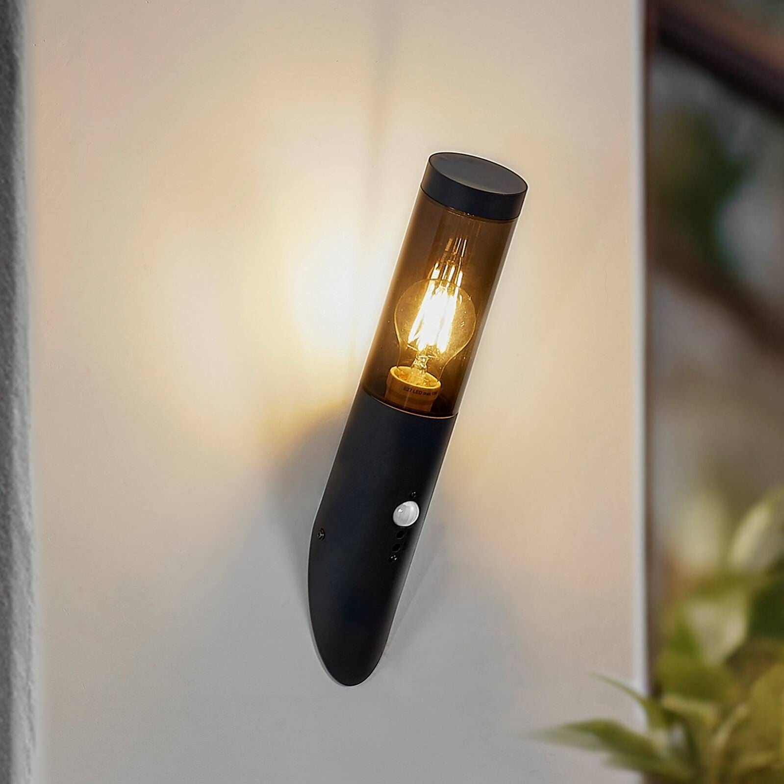 Outdoor wall light with motion sensor, black metal, cylindrical glass shade, emits warm light.