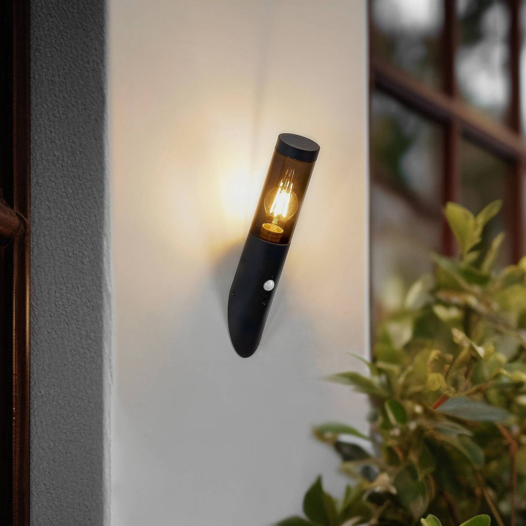 Black metal outdoor wall light with cylindrical glass cover and motion sensor, warmly glowing.