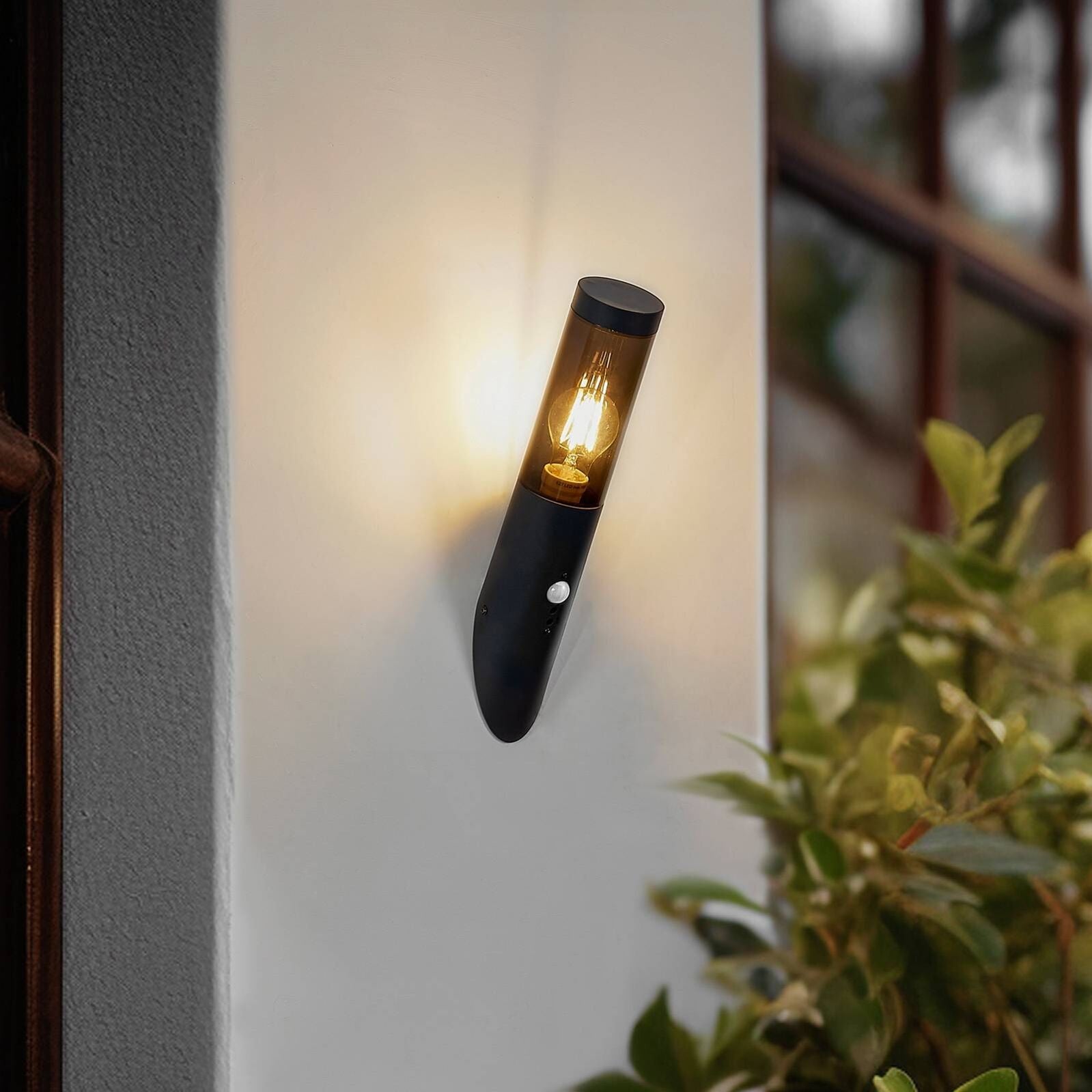 Black metal outdoor wall light with cylindrical glass cover and motion sensor, warmly glowing.